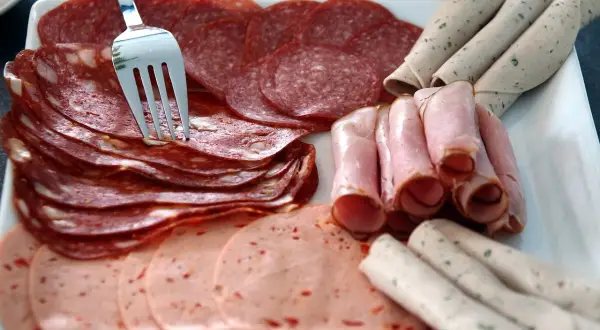 Eine Auswahl an Aufschnitt, darunter Salami und Schinken, liegt auf einer Platte. Eine Gabel steckt in den Scheiben, bereit zum Servieren oder zur Aufbewahrung.