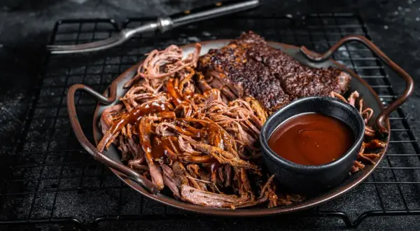 Saftiges pulled pork mit BBQ-Sauce und Beilagen auf einem Teller. Ein Gabel liegt daneben.