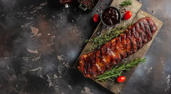 Saftige Rippchen, perfekt mariniert und gegrillt, liegen auf einem Holzbrett. Dazu gibt es BBQ-Sauce, Rosmarin und Tomaten.