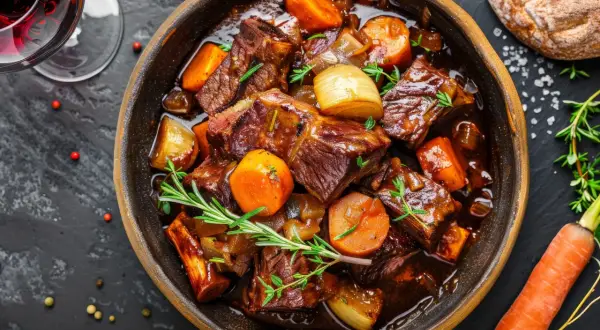 Herzhaftes Boeuf Bourguignon in einer Schüssel, mit Karotten, Zwiebeln und Rosmarin garniert. Daneben ein Glas Rotwein und ein Brot.
