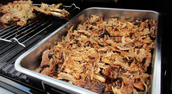 Saftiges pulled pork liegt in einer Metallschale auf dem Gasgrill. Daneben auf dem Rost ein Teil eines Rippchenbratens.