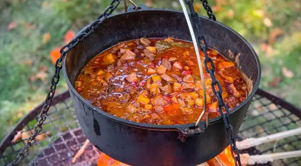 Herzhaftes Kesselgulasch-Rezept, das über offenem Feuer in einem Gusseisentopf köchelt. Fleischstücke, Kartoffeln und Paprika in einer würzigen Sauce.