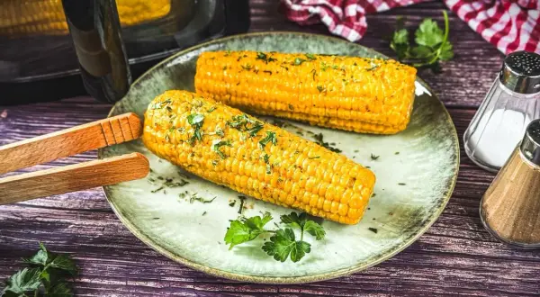 Zwei goldgelbe Maiskolben, frisch aus dem Airfryer, liegen auf einem Teller, garniert mit Kräutern. Perfekt für einen leckeren Snack!