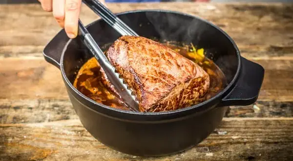 Ein saftiges Stück Fleisch wird mit einer Zange aus einem Topf gehoben, in dem es in einer dunklen Soße schmort. Das langsame Garen, das Schmoren, macht es zart.