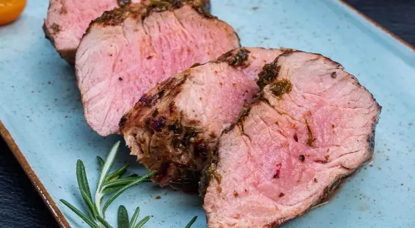Saftiger schweinelummer, perfekt gegrillt und mit Kräutern verfeinert, liegt auf einem blauen Teller. Ein Rosmarinzweig und eine halbe Tomate garnieren das Gericht.