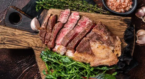 Perfekt gegartes Côte de Boeuf sous vide, in Scheiben geschnitten, mit frischem Grünzeug und Rosmarinzweig auf Holzbrett.