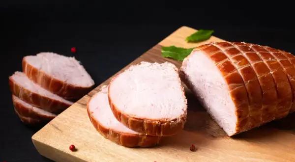 Ein aufgeschnittener Kassler-Braten mit perfekter Kerntemperatur, garniert mit Pfefferkörnern und Minze auf einem Holzbrett.