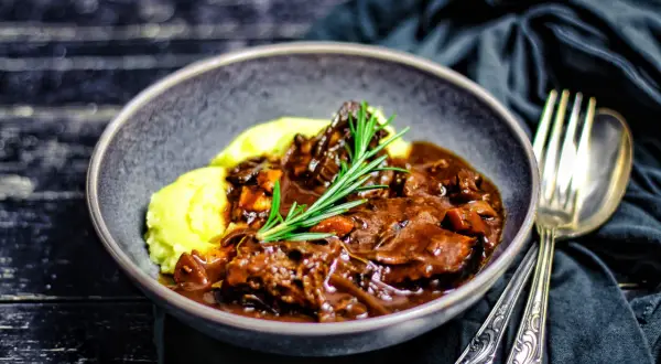 Ein Teller mit cremigem Kartoffelpüree und einem reichhaltigen Stracotto di manzo, garniert mit Rosmarin. Daneben liegen silberne Gabel und Löffel.