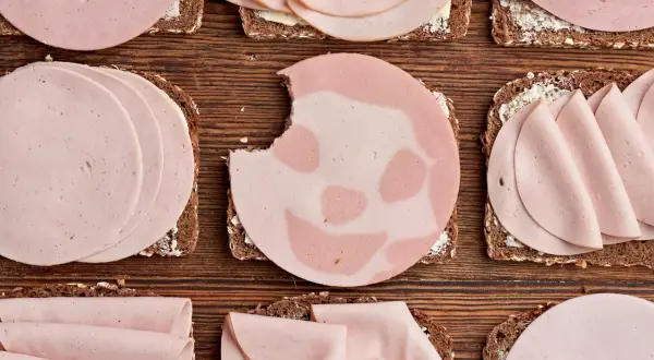 Biss in Lyoner: Welche Wurst ist gesund? Mehrere Brotscheiben mit Wurst belegt, liegen auf Holzuntergrund.
