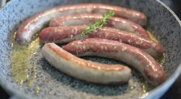 Fünf rohe Bratwürste brutzeln in einer Pfanne mit Butter. Ein Zweig Thymian liegt auf den Würsten, ein Hinweis auf ein köstliches wildbratwurst rezept.