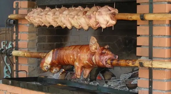 Ein Spanferkel grillen über offenem Feuer. Darüber hängen mehrere Hähnchen am Spieß.