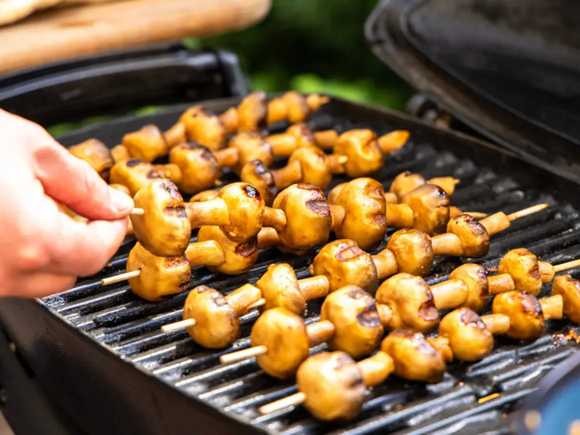 Leckere Champignons grillen auf dem Rost. Eine Hand steckt einen weiteren Spieß mit Pilzen auf den Grill.