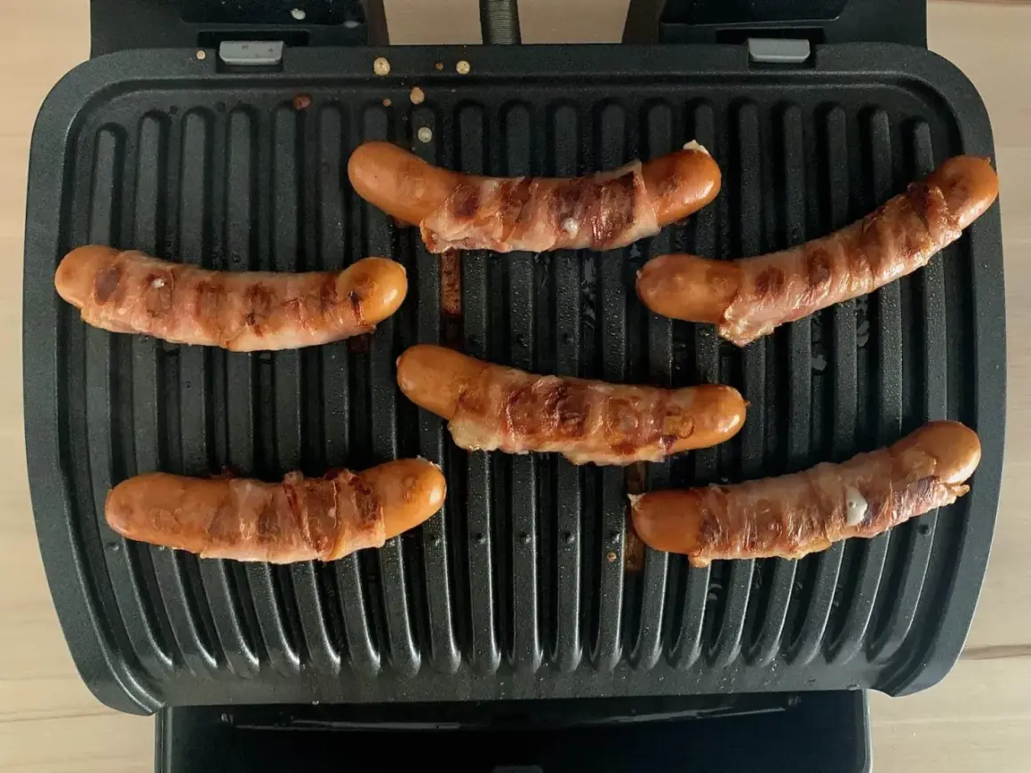 Sechs **Berner Würste** brutzeln auf einem Kontaktgrill. Die Würste sind mit Speck umwickelt und werden goldbraun.