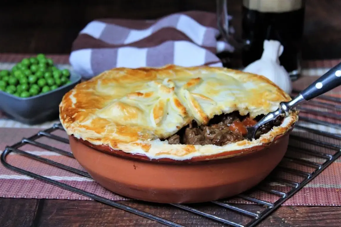 Herzhaftes Steak Pie mit goldbrauner Kruste, dane eine Schüssel Erbsen und ein Glas dunkles Bier.