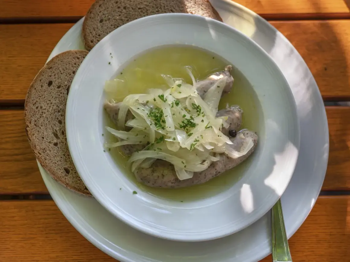 Drei **blaue Zipfel** in einer Schüssel mit Zwiebeln und Brühe, dazu zwei Scheiben dunkles Brot auf einem Holztisch.