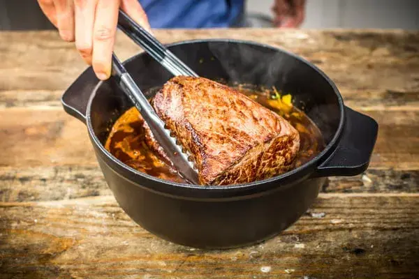 Ein saftiges Stück Fleisch wird mit einer Zange aus einem Topf gehoben, in dem es in einer dunklen Soße schmort. Das langsame Garen, das Schmoren, macht es zart.