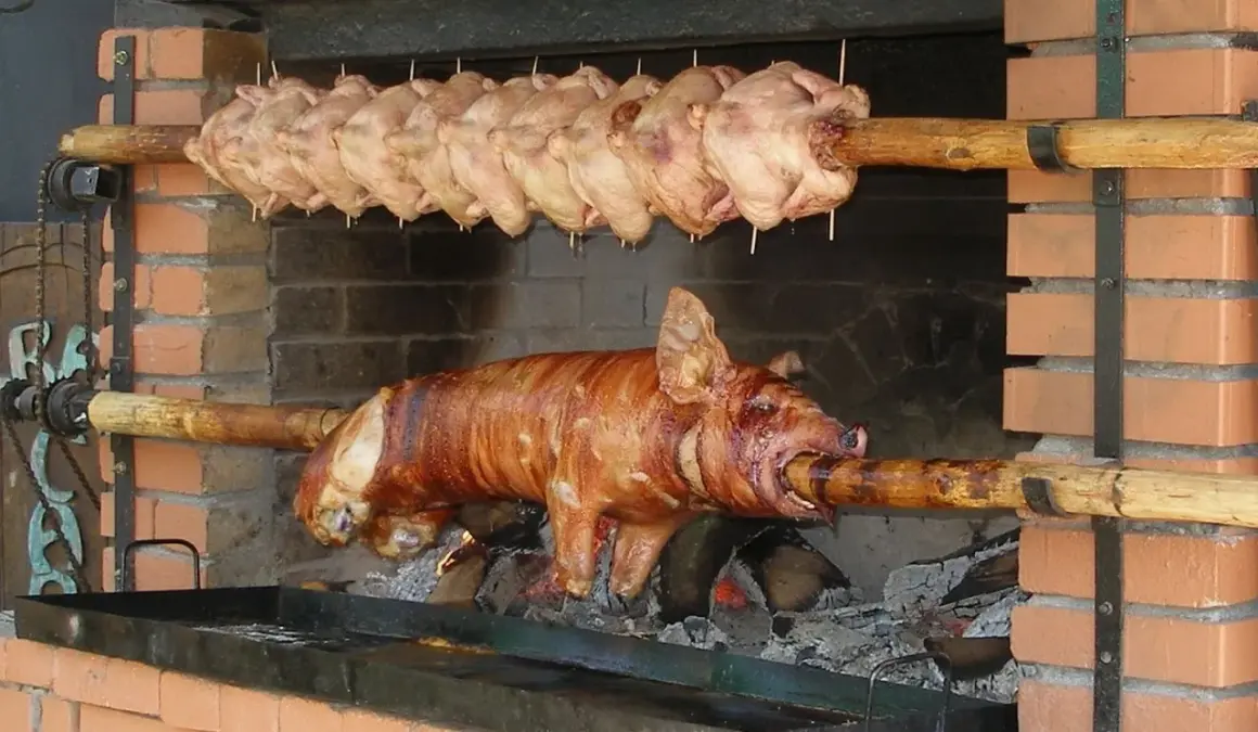 Ein Spanferkel grillen über offenem Feuer. Darüber hängen mehrere Hähnchen am Spieß.