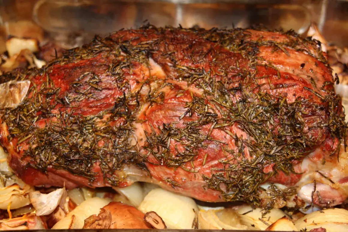 Saftige Lammkeule mit Rosmarin, bereit zum Grillen, liegt auf gerösteten Zwiebeln und Knoblauch.