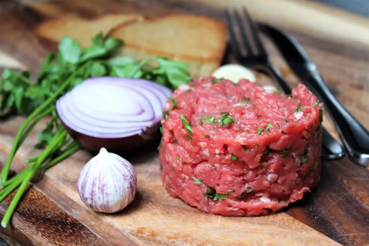 Frisch zubereitetes Beef Tatar mit Zwiebeln, Knoblauch und Petersilie auf einem Holzbrett. Dazu geröstetes Brot und Besteck.