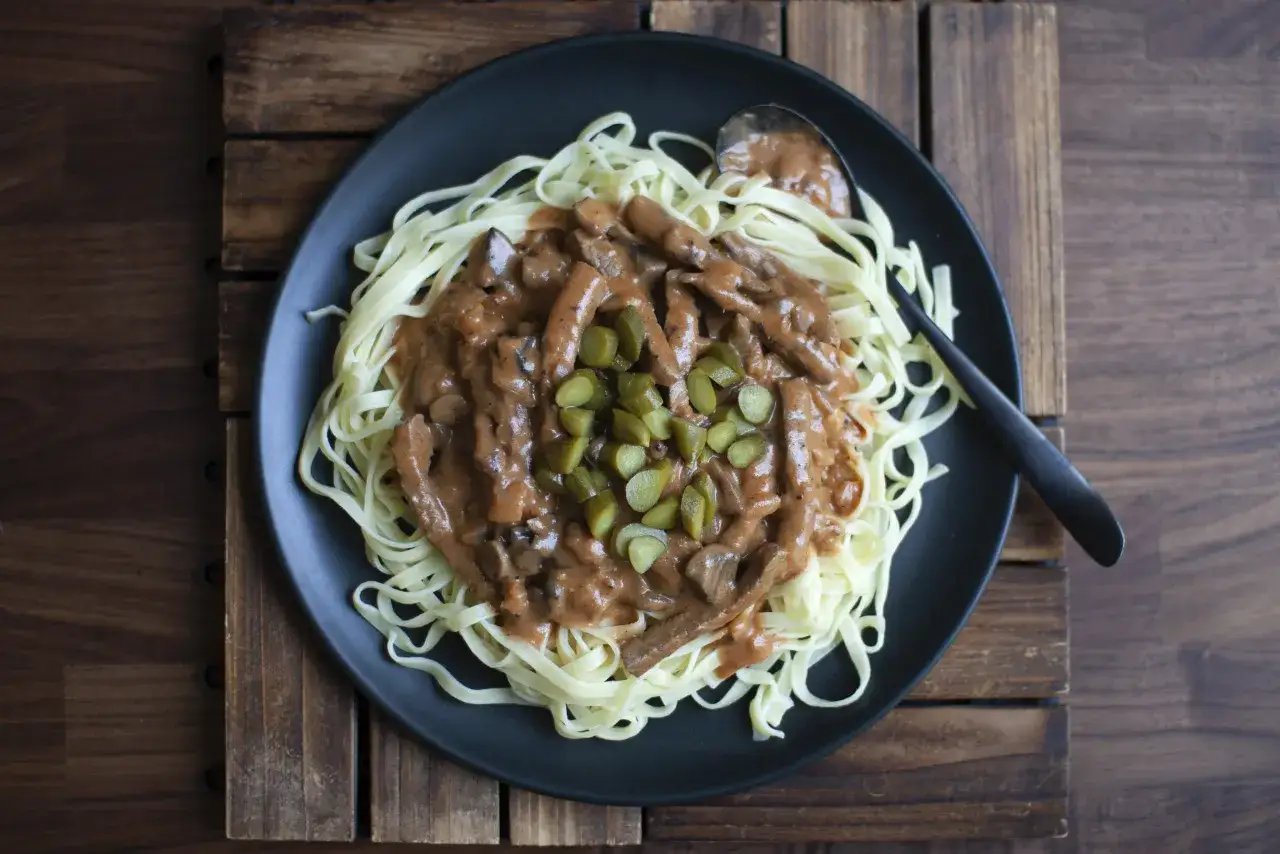 Ein Teller mit Bandnudeln und cremigem Boeuf Stroganoff Rezept, garniert mit Gewürzgurken. Ein Löffel liegt bereit.