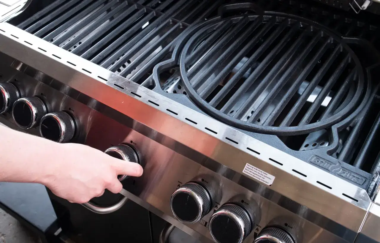 Hand dreht Regler am Gasgrill zum Einbrennen. Neue Grillroste warten auf ihren ersten Einsatz.