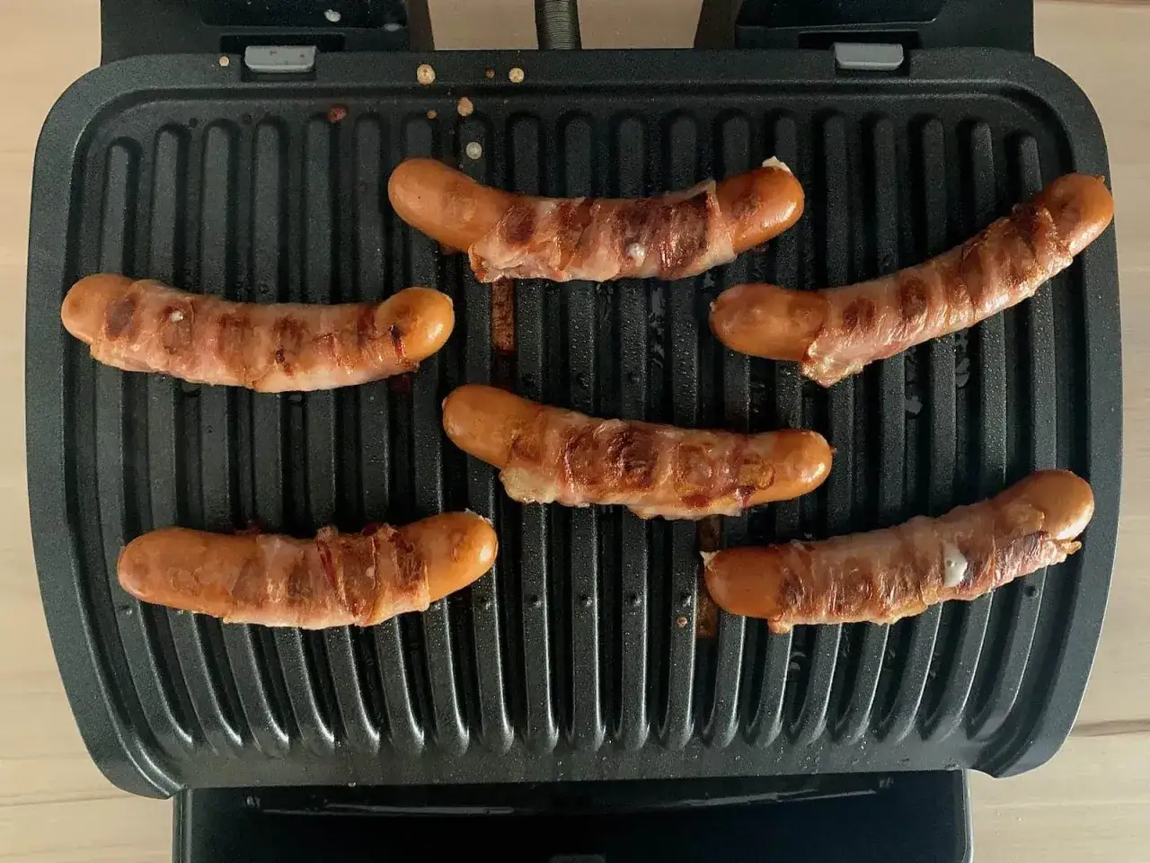 Sechs Berner Würste brutzeln auf einem Kontaktgrill, umhüllt von knusprigem Speck.