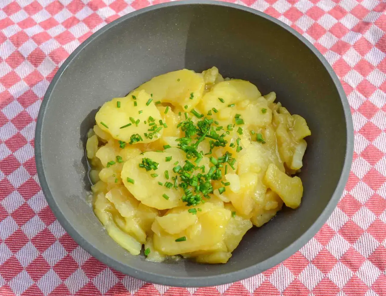 Omas Kartoffelsalat, ein Klassiker mit zarten Kartoffelscheiben und frischem Schnittlauch, serviert in einer grauen Schüssel auf rot-weiß kariertem Tuch.