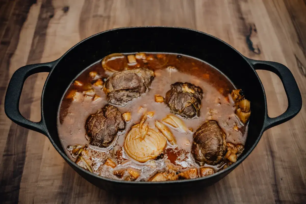 Ein Gusseisentopf mit Rouladen, Zwiebeln und Gemüse in einer dunklen Soße. Das langsame Garen, das Schmoren, macht das Fleisch zart und die Soße geschmacksintensiv.