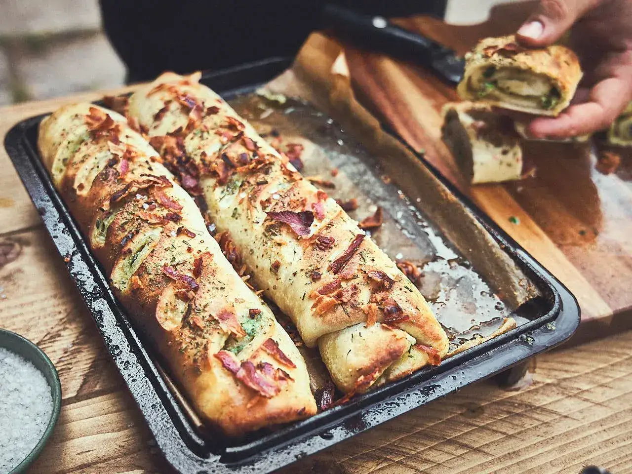 Zwei goldbraune Baguettes zum Grillen, belegt mit Speck und Kräutern, liegen auf einem Blech. Daneben ein Holzbrett mit angeschnittenen Stücken.