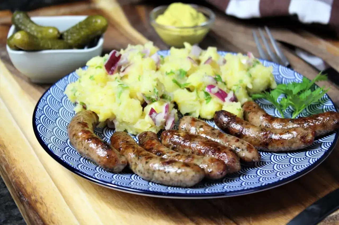 Bratwurst selber machen: Gebratene Würstchen mit Kartoffelsalat, Gewürzgurken und Senf auf blau-weißem Teller.