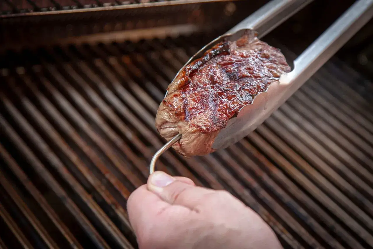 Ein Steak wird mit einer Zange &uuml;ber einem Grill gehalten, um die Kerntemperatur Rind zu pr&uuml;fen.