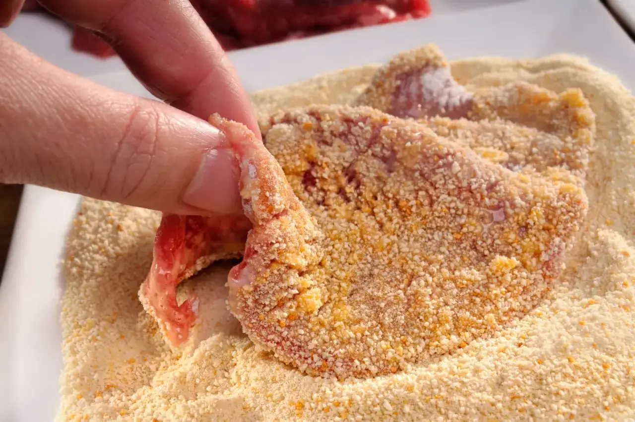 Eine Hand wendet ein rohes Schnitzel in Paniermehl. Der Prozess des Schnitzel panieren ist im Gange.