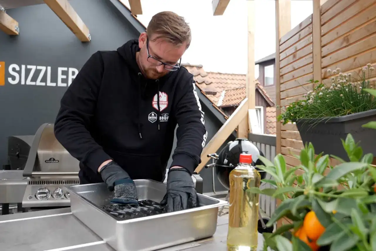 Mann mit Brille reinigt mit B&uuml;rste den Grillrost in einer Wanne. So wird der Grillrost reinigen zum Kinderspiel.