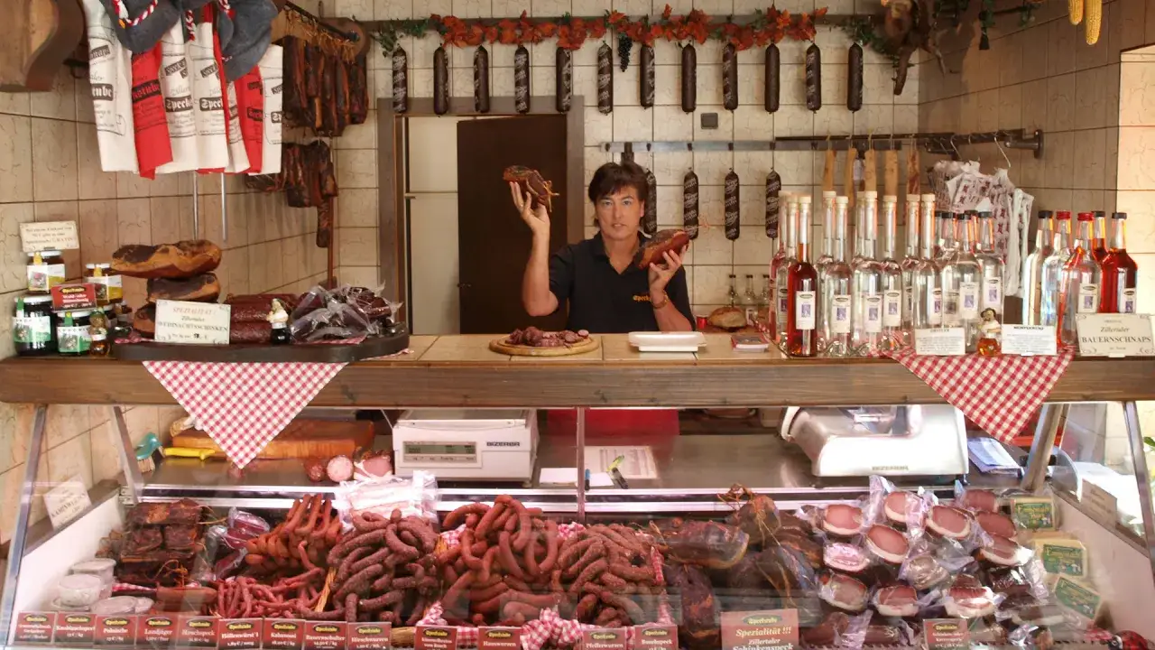 Fleischerei-Theke mit vielerlei Wurst und Schinken, ideal zum wurst aufbewahren. Eine Verkäuferin hält ein Stück Fleisch hoch.