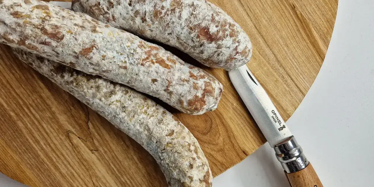 Drei luftgetrocknete Salamis liegen auf einem Holzbrett, daneben ein Messer. Man fragt sich, ob die Salami schlecht werden kann.