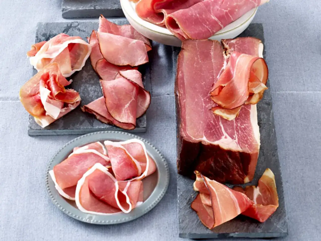 Verschiedene Scheiben und ein Stück Schinken vom Schwein, appetitlich auf grauen Platten und einem Teller arrangiert.