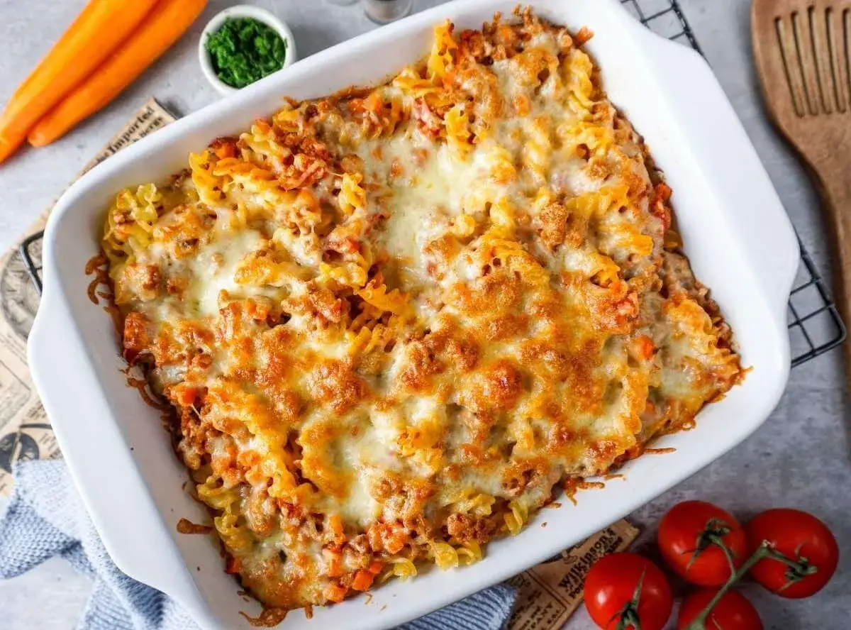 Ein köstlicher Nudel Hackfleisch Auflauf mit geschmolzenem Käse, serviert in einer weißen Auflaufform. Daneben liegen Karotten und Tomaten.