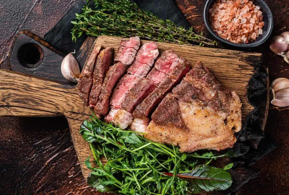 Perfekt gegartes C&ocirc;te de Boeuf sous vide, in Scheiben geschnitten, mit frischem Gr&uuml;nzeug und Rosmarinzweig auf Holzbrett.