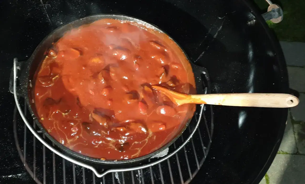 Currywurst in einer roten Sauce köchelt im Dutch Oven auf dem Grill. Ein Holzlöffel rührt um.