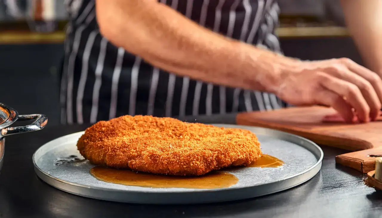 Ein goldbraunes Schnitzel liegt auf einem Teller mit Sauce. Ein Koch bereitet im Hintergrund weitere Zutaten vor, vielleicht zum Schnitzel klopfen.