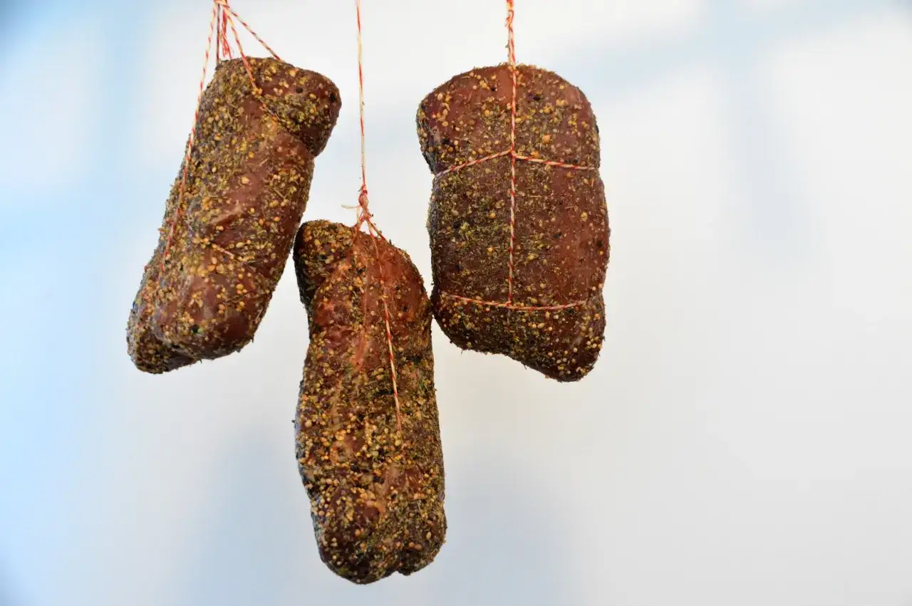 Drei geräucherte Fleischstücke, umhüllt von Kräutern und Gewürzen, hängen an Schnüren. Sie zeigen, woraus Lachsschinken besteht: aus sorgfältig gewürztem und gereiftem Fleisch.