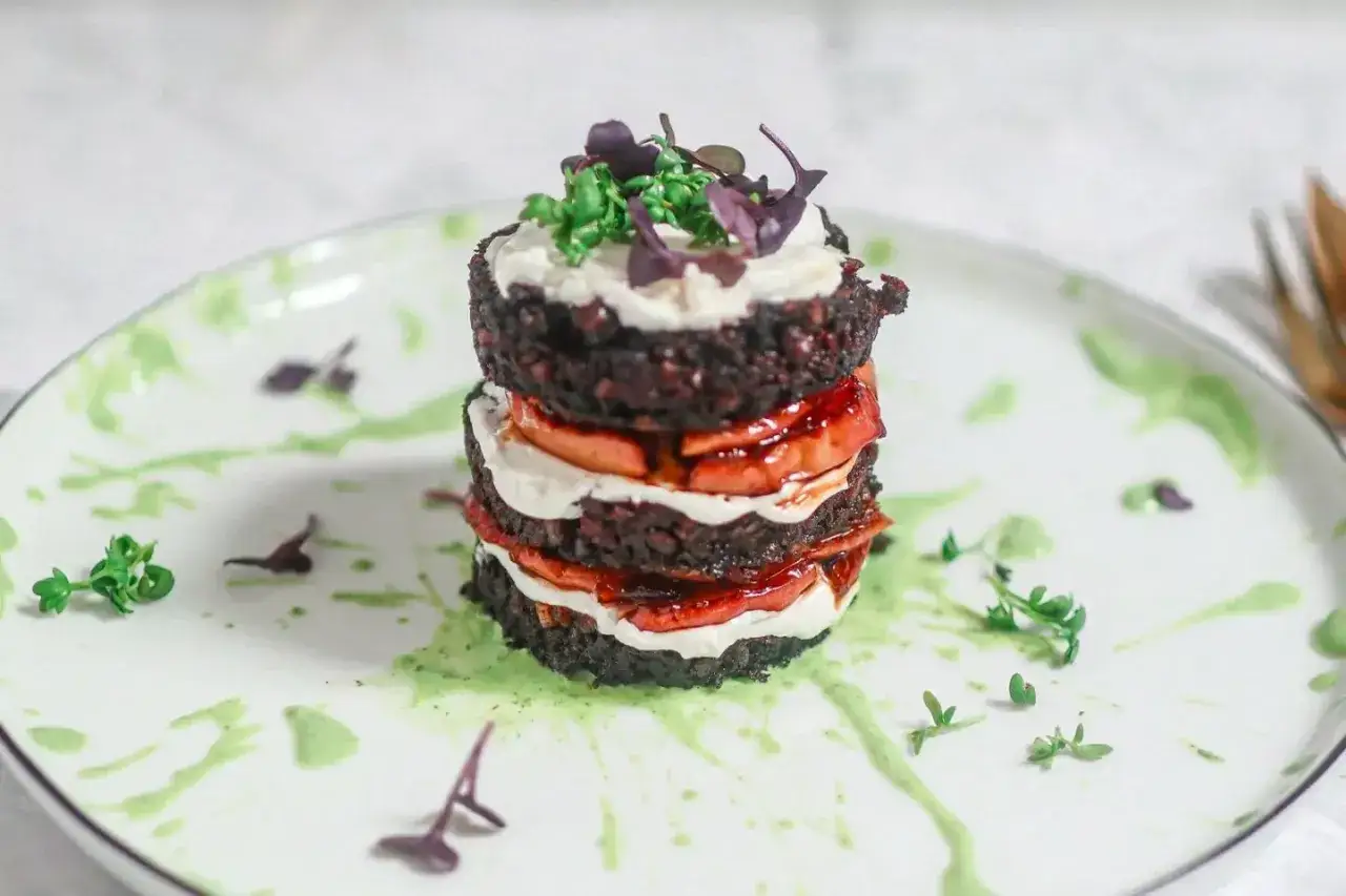 Ein gestapeltes Gericht mit dunklen Scheiben, die an Black Pudding erinnern, dazwischen cremige Füllung und rote Fruchtsauce.