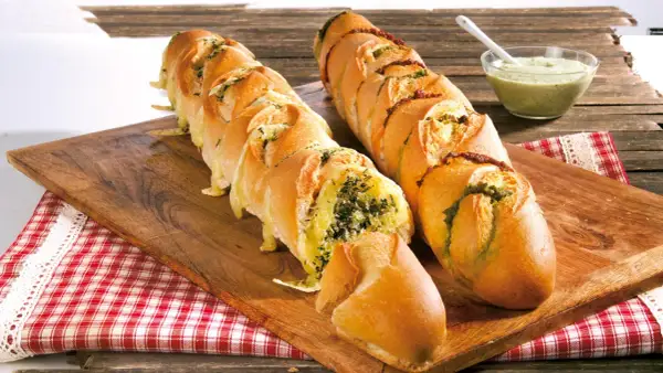 Zwei gefüllte Baguettes zum Grillen, eines mit Kräutern und Käse, das andere mit Pesto und Tomaten, daneben eine Schüssel Dip.