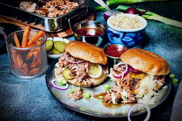 Zwei saftige Pulled Pork Burger, zubereitet im Sous Vide Verfahren, mit Krautsalat, Gurken und roten Zwiebeln. Dazu Pommes und BBQ-Sauce.