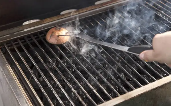 Eine Zwiebel wird mit einer Zange auf dem Grillrost platziert, um den Grill ausbrennen zu lassen. Rauch steigt auf.