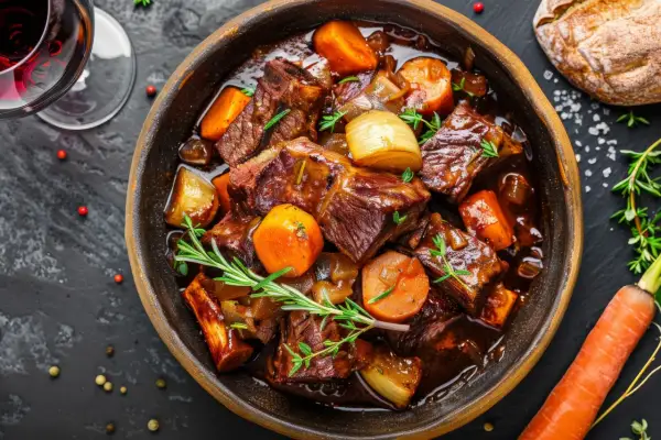 Herzhaftes Boeuf Bourguignon in einer Schüssel, mit Karotten, Zwiebeln und Rosmarin garniert. Daneben ein Glas Rotwein und ein Brot.