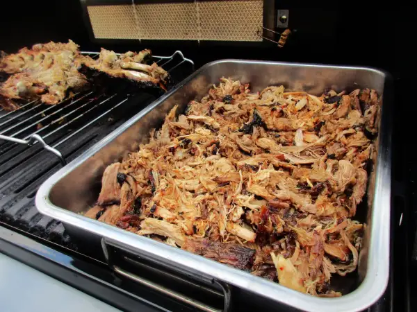 Saftiges pulled pork liegt in einer Metallschale auf dem Gasgrill. Daneben auf dem Rost ein Teil eines Rippchenbratens.