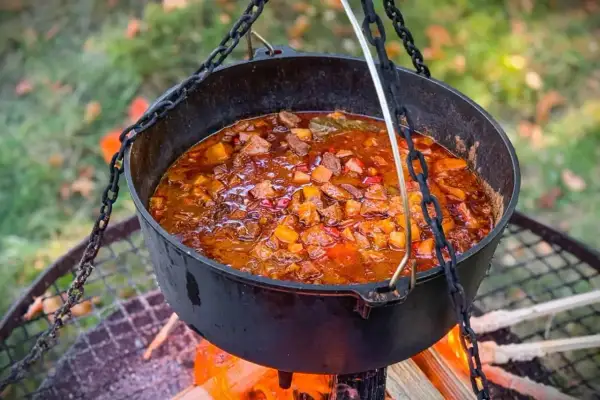 Herzhaftes Kesselgulasch-Rezept, das über offenem Feuer in einem Gusseisentopf köchelt. Fleischstücke, Kartoffeln und Paprika in einer würzigen Sauce.