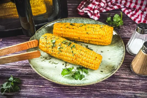 Zwei goldgelbe Maiskolben, frisch aus dem Airfryer, liegen auf einem Teller, garniert mit Kräutern. Perfekt für einen leckeren Snack!