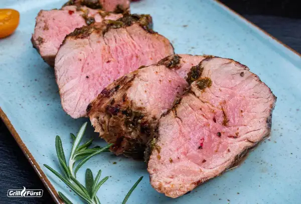 Saftiger schweinelummer, perfekt gegrillt und mit Kräutern verfeinert, liegt auf einem blauen Teller. Ein Rosmarinzweig und eine halbe Tomate garnieren das Gericht.